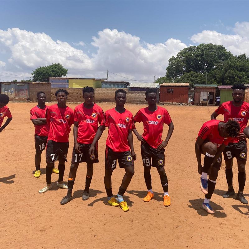A youth team in red kit lined up on a bare earth pitch beside township housing