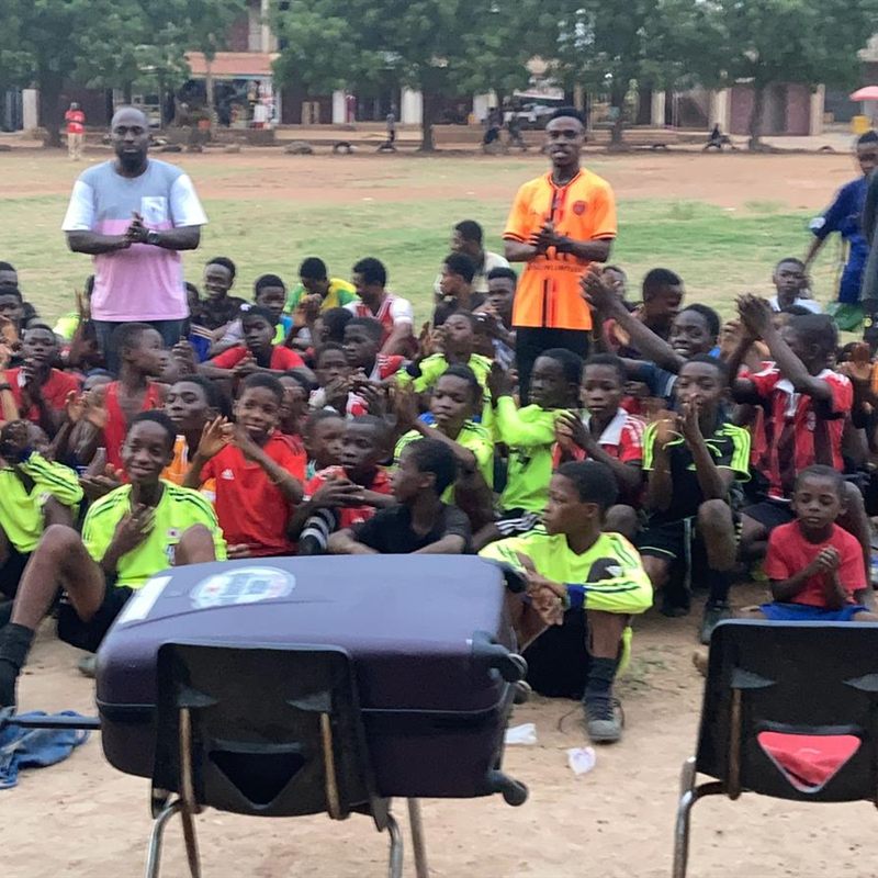 Dozens of children seated on the field with two coaches and a case of supplies