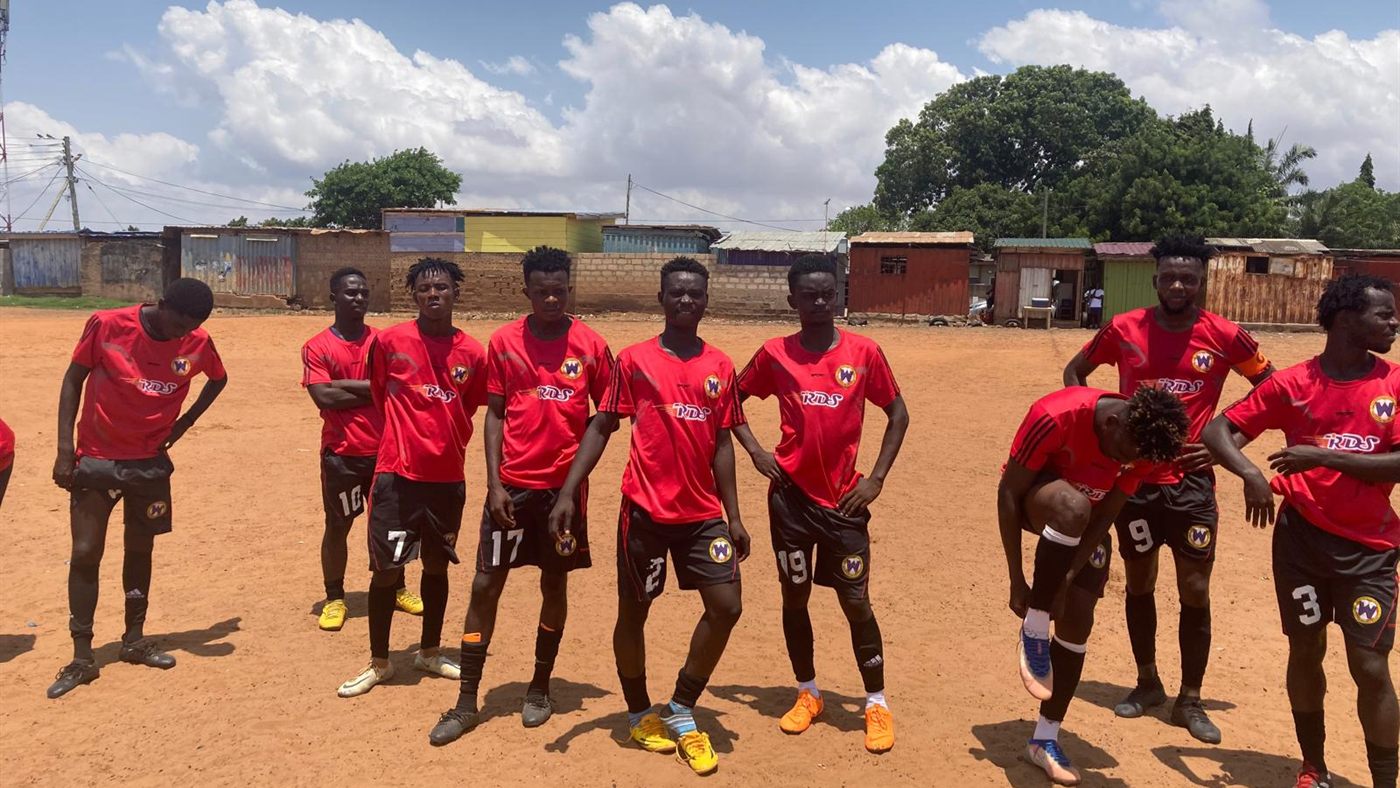 A youth team in red kit lined up on a dirt pitch in a Ghanaian township